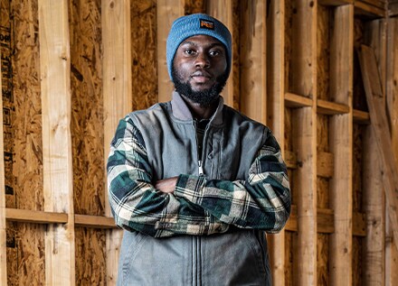 A man in a blue winter beanie, flannel shirt and grey vest, against the unfinished framed wall of a new construction house.