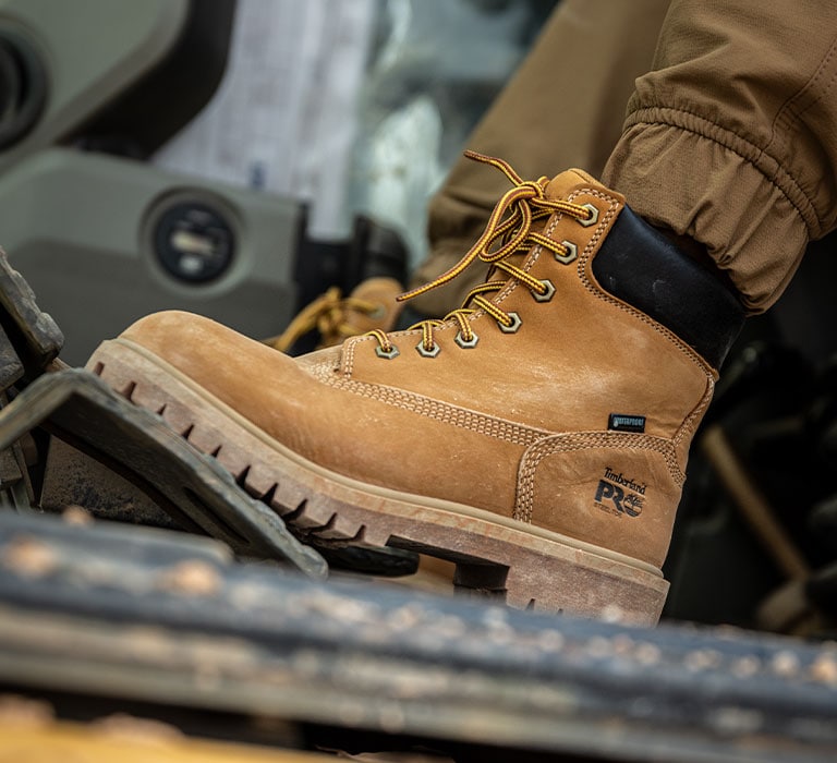 Close-up image of a Timberland wheat colored work boot with black collar and scuffed outsole. The person wearing the boot is using it to apply pressure to the gas pedal of a construction vehicle.