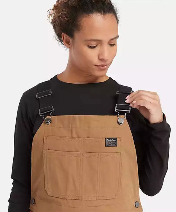 Image of a woman wearing a black long sleeve t-shirt and brown overalls, looking down at the overalls and holding the strap with one hand.