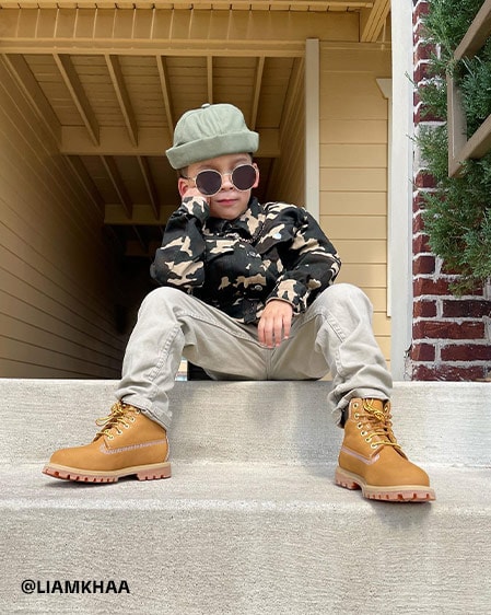 Image of a young boy showing some 'tude, sitting on the front stoop of a brick and yellow house, wearing an off-white hat and pants, black and white jacket, round sunglasses and classic wheat Timberland boots.