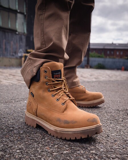 Image of a pair of wheat Timberland PRO boots being worn from the knee down by a man standing on gravel. His pants are brown and bunched around the boot collars, and the boots themselves are a bit scuffed up with wear and tear.