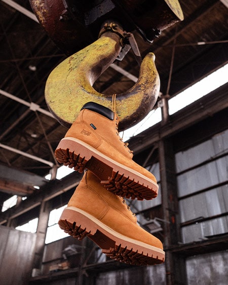 Image of a pair of wheat Timberland PRO workboots, laces suspended from a yellow crane hook that's coming down from the ceiling of a warehouse