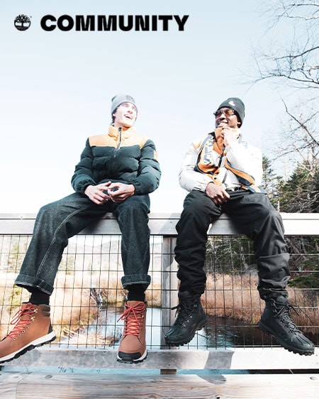 Image of two young men sitting on a bridge over a small stream, laughing and wearing Timberland hats, puffy coats, black pants and laced boots.