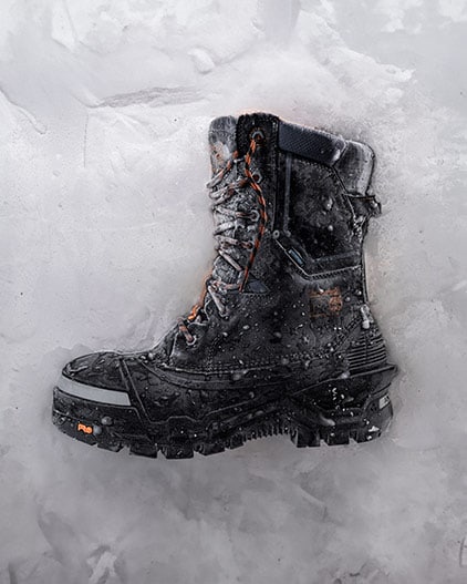 Image of a tall black Timberland PRO work boot that's half-encased in snow, on its side, with ice and snow encroaching on the leather and laces.