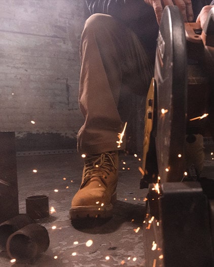 Image of a man sitting in a dark room with concrete brick walls, using some soft of industrial saw that's emitting sparks. The image is focused around the man's wheat Timberland lace-up work boot, which is aimed toward the camera.