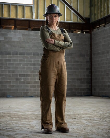 Image of a woman in brown braids standing in a warehouse on concrete, wearing a black hard hat and brown Timberland work overalls, arms crossed and staring at the camera.