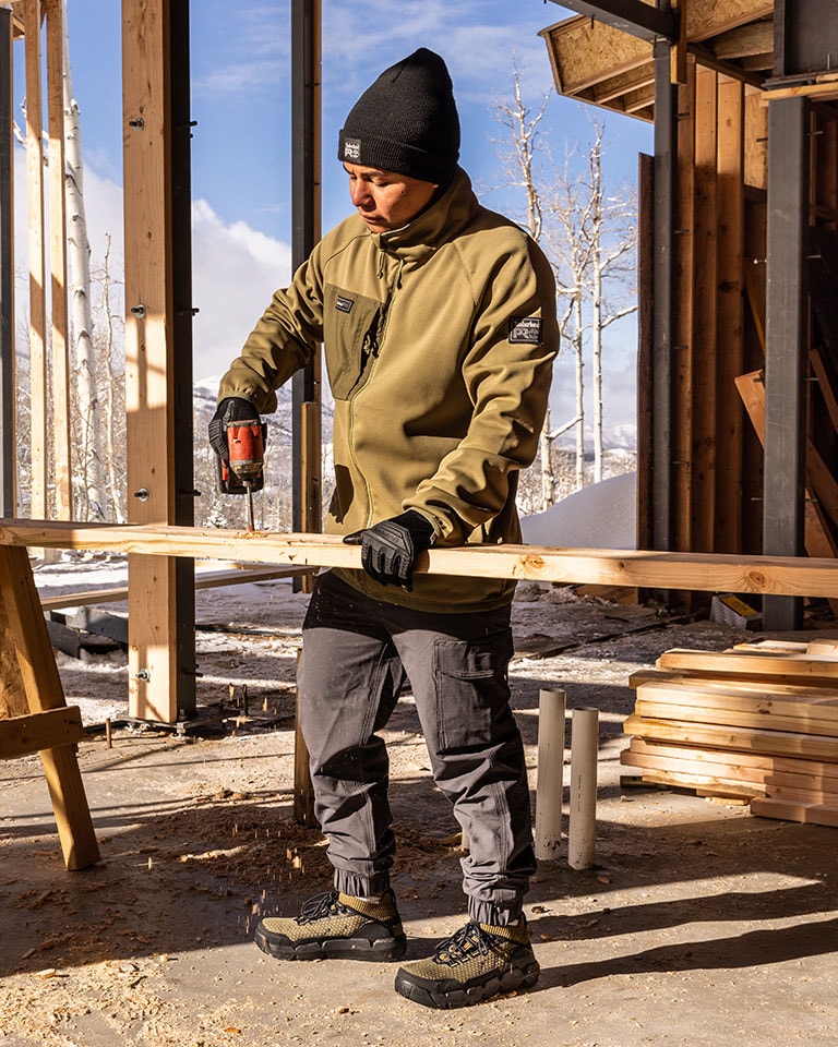 Image of a man standing outside at a construction site with blue skies, snowcapped mountains and bare trees in the background. He's wearing a black Timberland PRO beanie, black work gloves and a tan Timberland PRO work jacket as he uses a drill to drill into a piece of lumber.