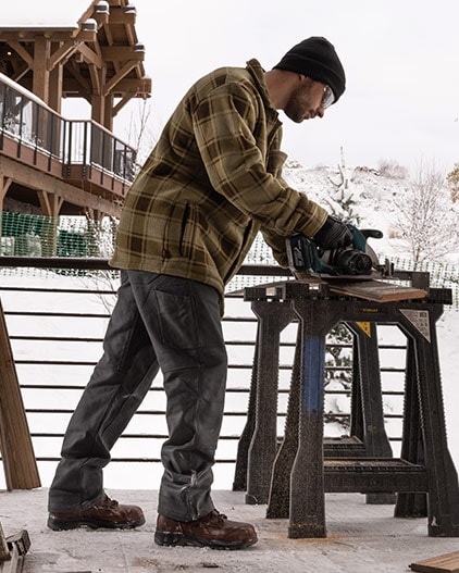 A man wood-working outside in front of a snowy back drop, he is wearing a brown flannel, grey Timberland PRO work pants and brown work boots. 