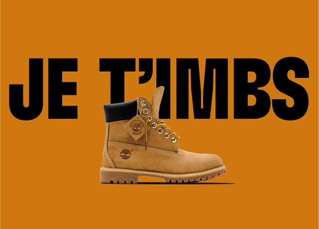 Timberland Premium Yellow boot unlaced and floating on an orange background with the text JE T'IMBS behind it.