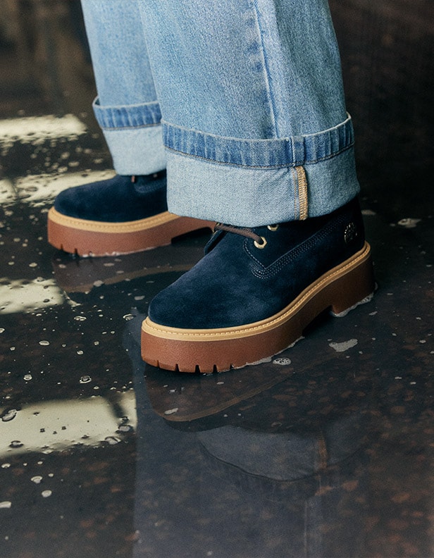 Image of a closeup from the calf down of a person wearing Timberland Indigo Suede dark blue boots and baggy, light wash denim with the cuffs rolled up dramatically to show off the boots' gold hardware.