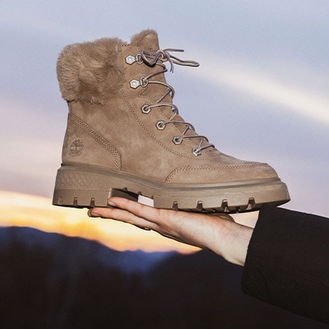 Woman holding a Timberland boot with the mountains and sky in the background.