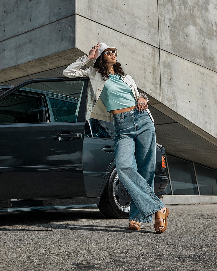 Image of a woman outside near a concrete building, leaning against a black car with the door open, wearing jeans, a light blue cropped t-shirt and white jacket and bucket hat, and brown leather Timberland slide sandals.