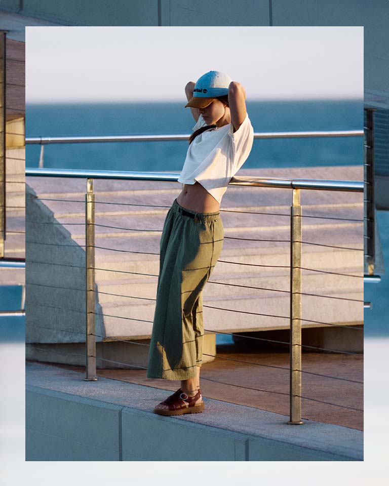 Image of a woman on a pier with the ocean behind her, wearing a white t-shirt, baseball cap and green flowy pants, with brown strappy sandals.