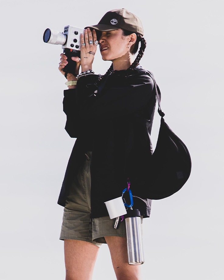 Woman standing against a white background, looking through a camera, wearing a black Timberland jacket, black Timberland visor and shorts, with a black sling bag and silver water bottle clipped to it.