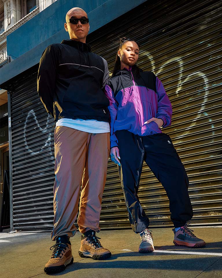 Image of a woman from the waist up, pictured from below, standing on a city street with blue sky behind her, holding open a purple windbreaker to reveal a black hoodie with TIMBERLAND in white letters. Image of a young man and woman standing on a city street side by side, wearing Timberland hikers, joggers and windbreakers. Image of a man on a city street, wearing sunglasses and a black windbreaker and leaning against a car.