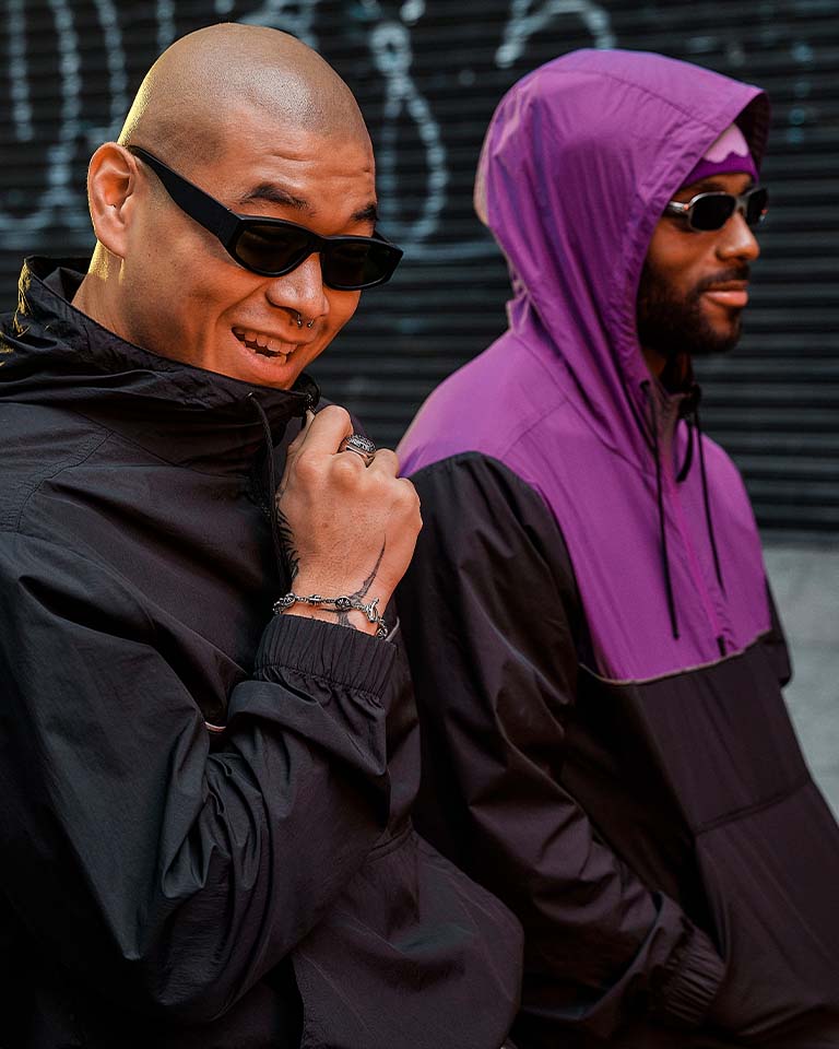Image of a man leaning back against a black car tailgate, wearing a black and purple windbreaker with the purple hood up. Image of two men walking down a city street with graffiti in the background, from the waist up, wearing black and purple windbreakers and sunglasses. Image of a man in sunglasses leaning against a car on a city street wearing a black windbreaker.