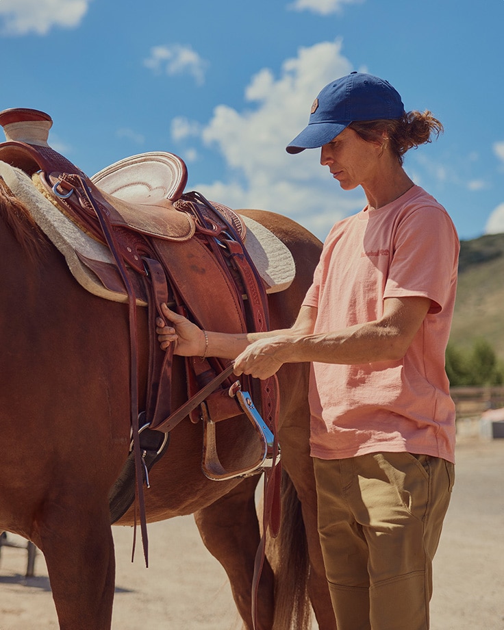 image of a woman outside wearing a light salmon pink t-shirt and a blue cap standing next to a horse holding the reins