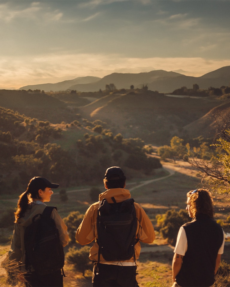 Image of three people in their 20s standing on top of a hill at golden hour, facing away from the camera. Two people are wearing wearing Timberland hiking jackets, backpacks and baseball caps, and one is wearing a vest over a white t-shirt with sunglasses on top of their head.