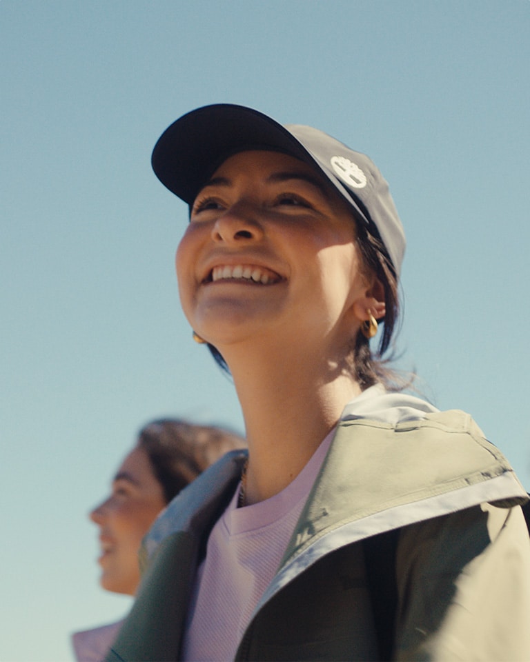 Close-up image of a woman in a Timberland blue baseball cap, taupe jacket and purple tee, from the chest up, smiling up into the blue sky.